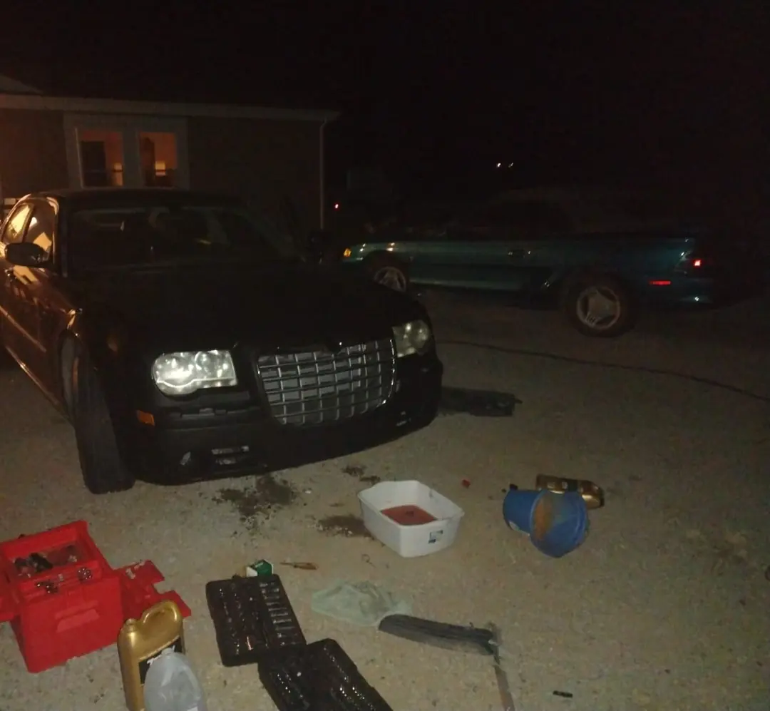 A black Chrysler 300 at night, toolboxes, a drain pan and oil spread on the gravel in front of it.
