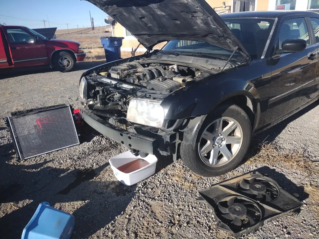 A black Chrysler 300 on a gravel lot with its front bumper off, the radiator and cooling fans on the ground beside it.