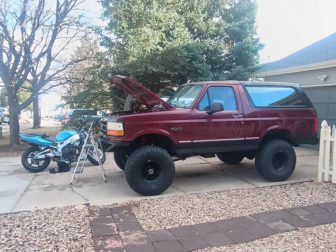 A red lifted Ford Bronco with its hood up in a driveway, a blue sport motorcycle on a stand beside it.