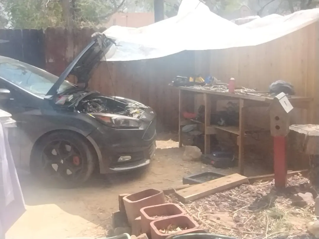 A black Ford Focus with its hood up in a backyard, a tarp rigged overhead for shade and a workbench of tools beside it.