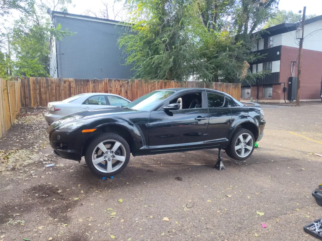A black Mazda RX-8 raised on jack stands in a gravel lot.