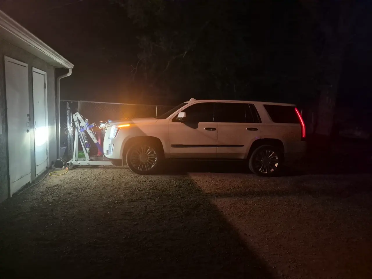 A white Cadillac Escalade in a gravel driveway at night, an engine hoist at its front end under a work light.
