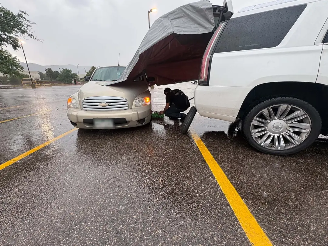 A mechanic crouched beside a car in a wet parking lot, working under an SUV's open rear hatch rigged as a rain shelter.