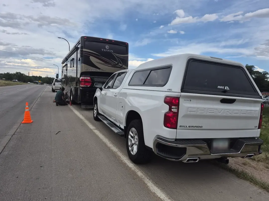 A motorhome stopped on a highway shoulder with a safety cone behind it, a mechanic kneeling at its rear wheel.
