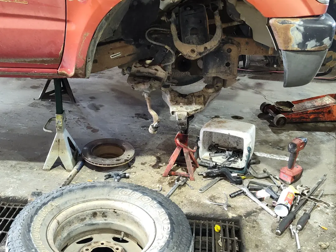 A red Toyota Tacoma on jack stands with its front wheel off, the brake caliper hanging free and the brake rotor on the floor beside it.